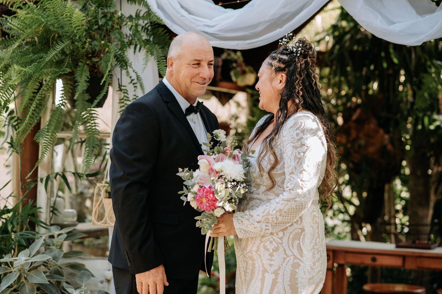 A wedding ceremony under a decorated arch with white draping and greenery.