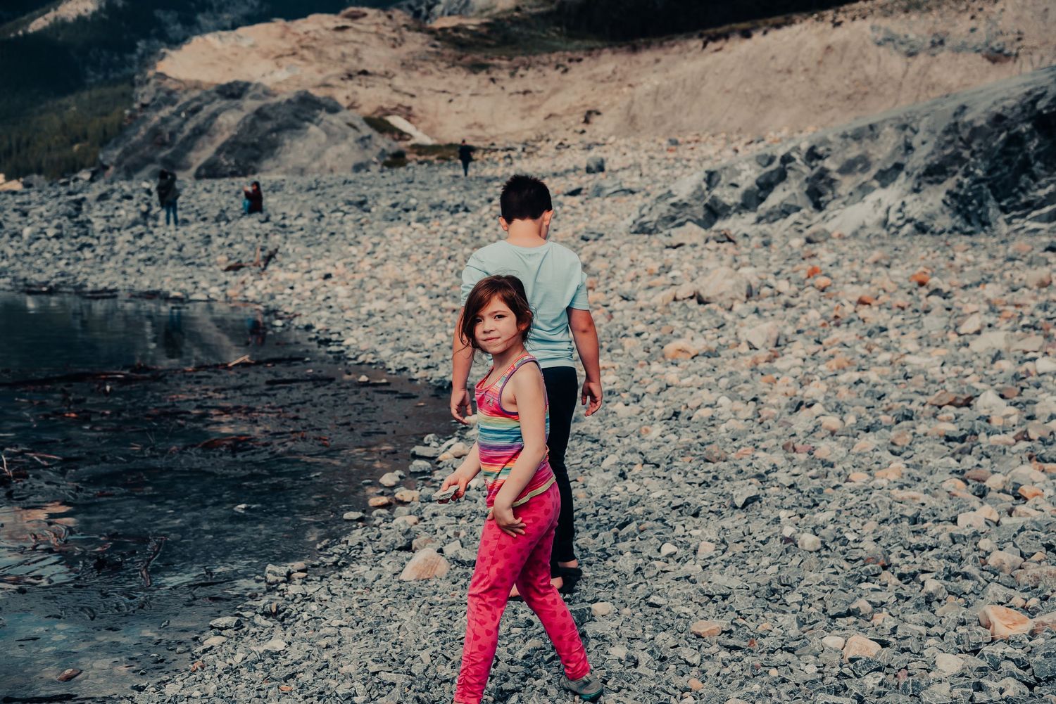 Two people exploring a rocky beach landscape against mountainous terrain.