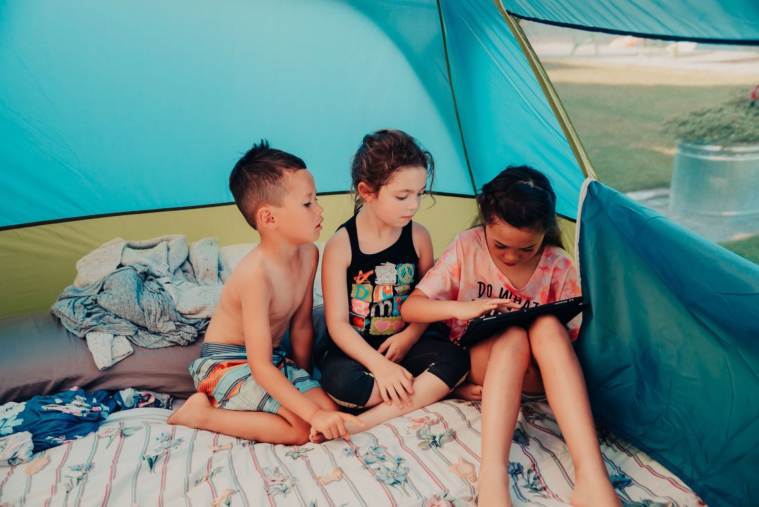 Three kids sitting together inside a blue tent during summer camping.