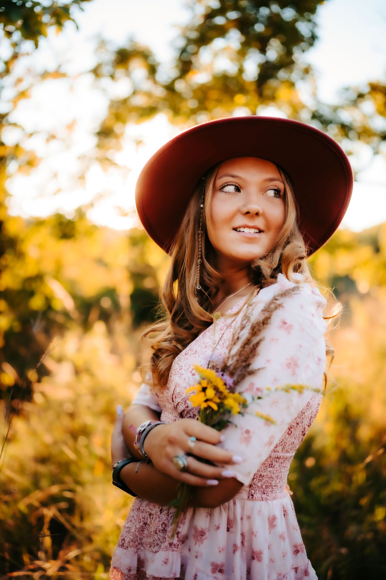 Woman in hat and floral dress holding wildflowers in a sun-drenched field.