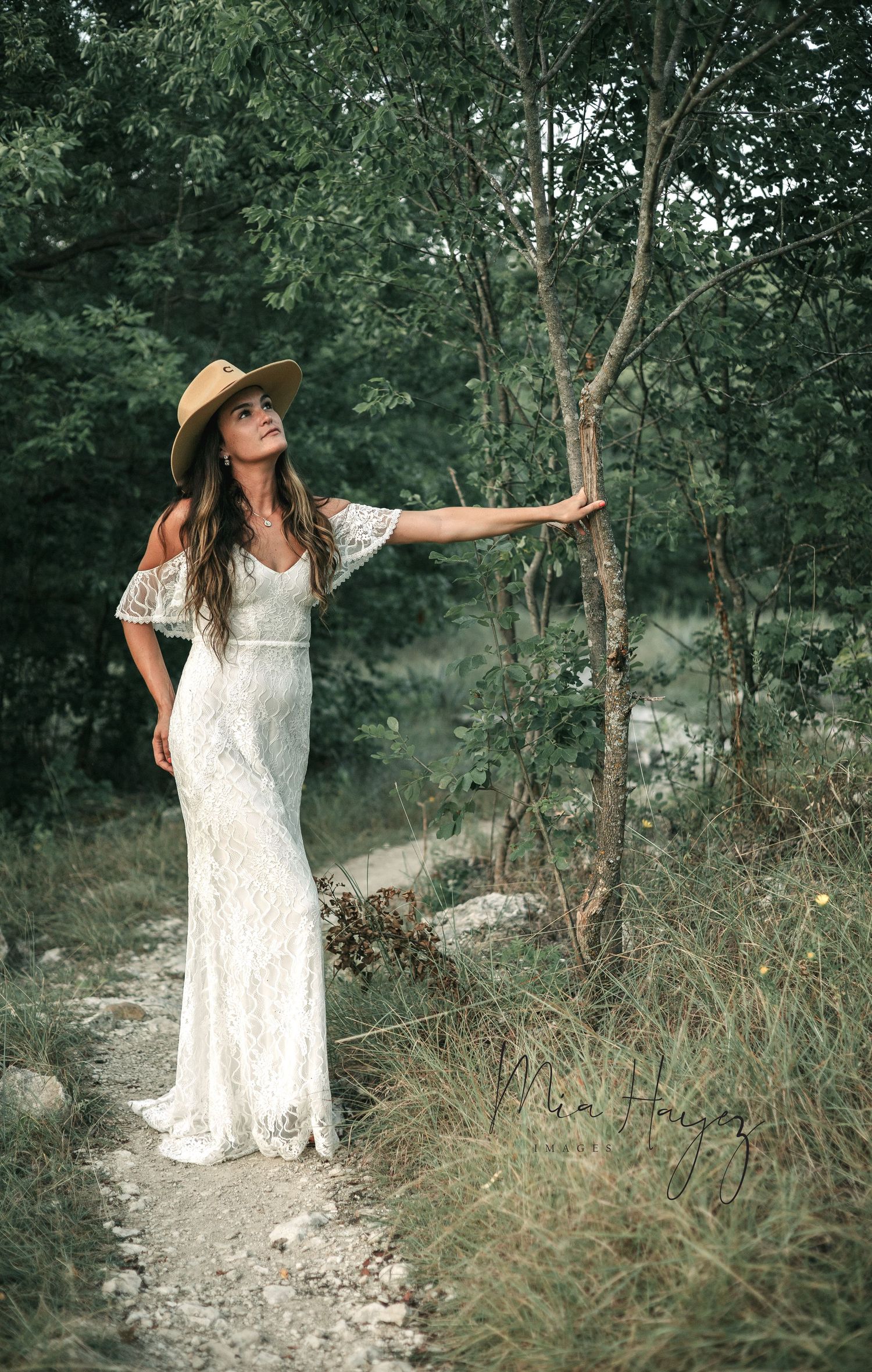 Woman in white lace dress poses gracefully in a wooded outdoor setting during a photoshoot.