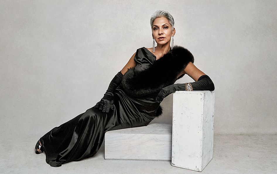 Elegant woman in a black satin gown with fur trim, posing dramatically against a gray background.