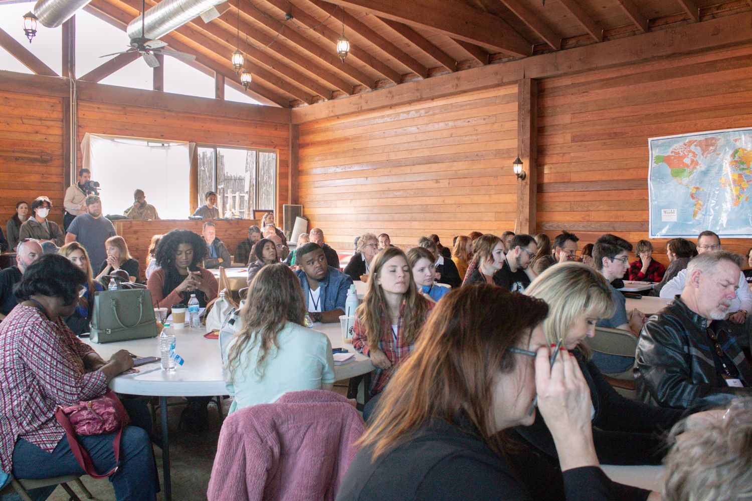 Large group of attendees seated at round tables inside a wooden lodge during a conference.