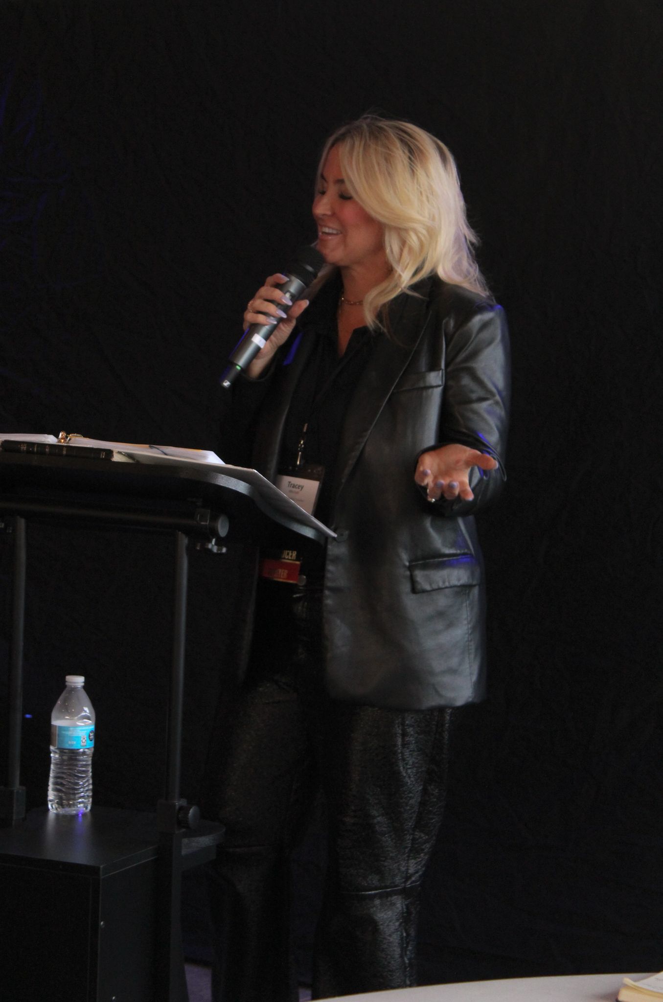 A blonde woman in a black leather blazer speaks into a microphone at a podium on stage.