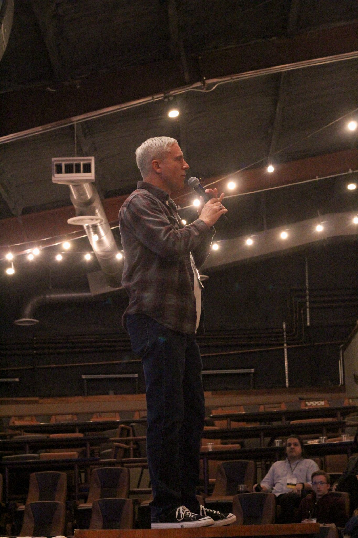 A gray-haired man in a plaid shirt speaks into a microphone on a dimly lit auditorium stage.