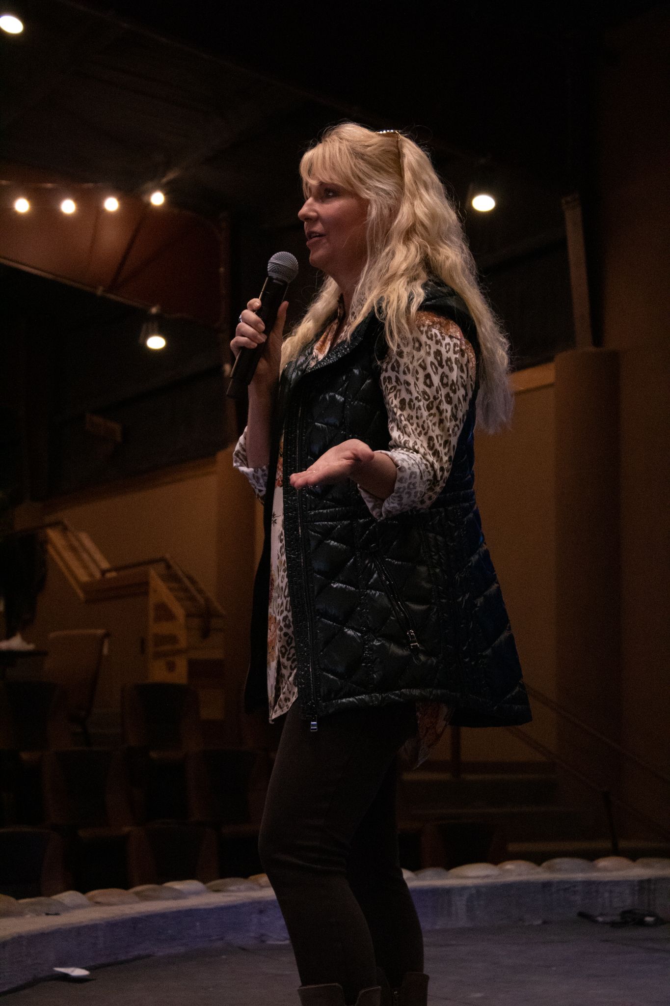 A blonde woman in a black quilted vest speaks into a microphone on a dimly lit stage.
