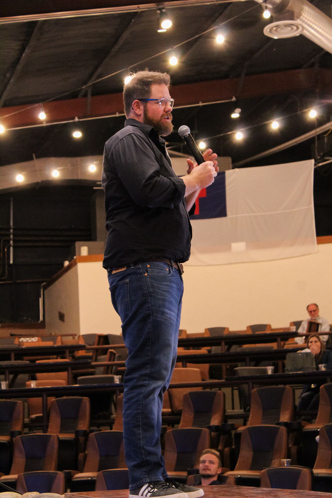 A bearded man in a black jacket speaks into a microphone on stage in a large auditorium.