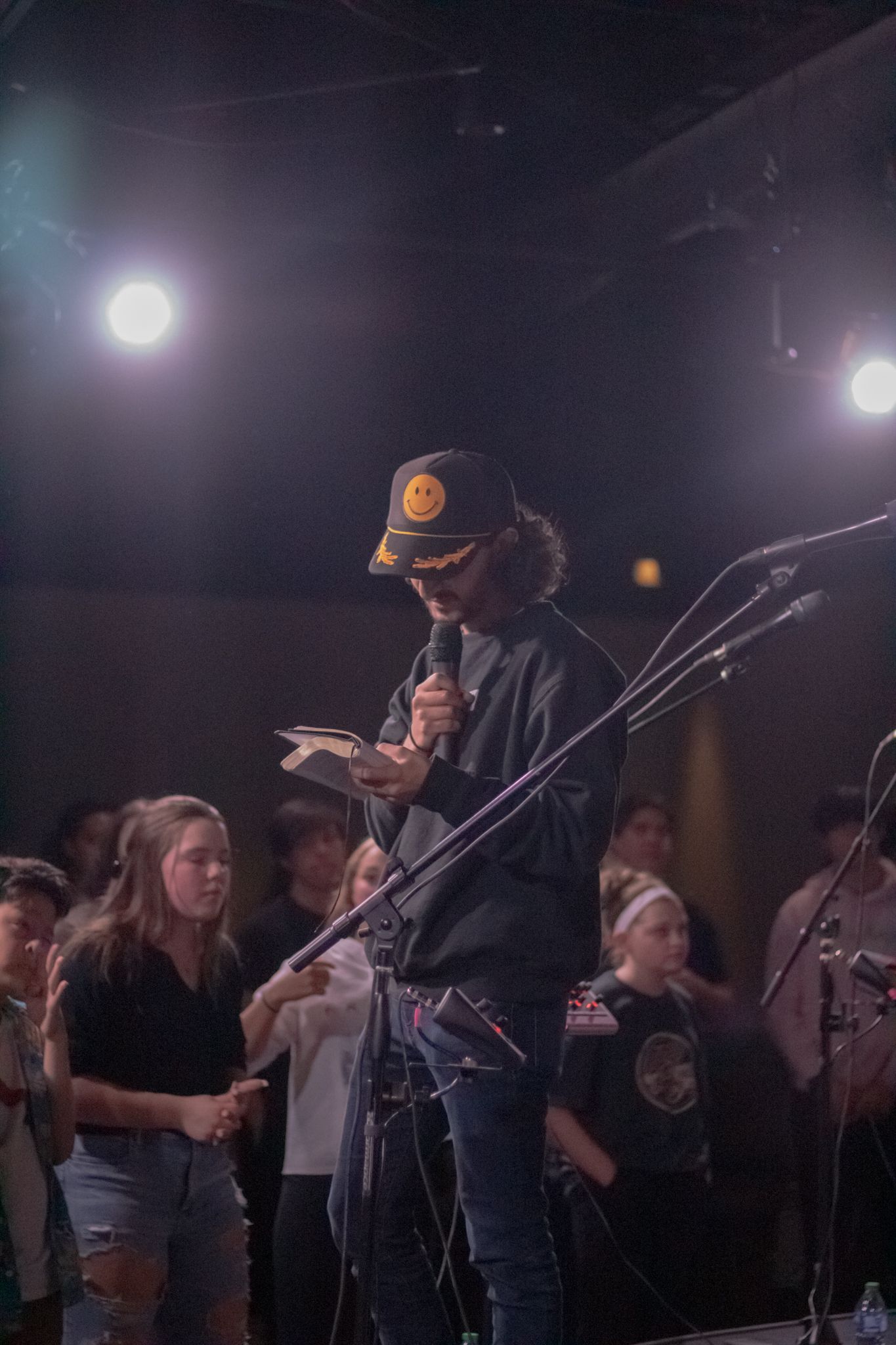 A young person in a smiley face cap reads from a book into a microphone on a dark stage.