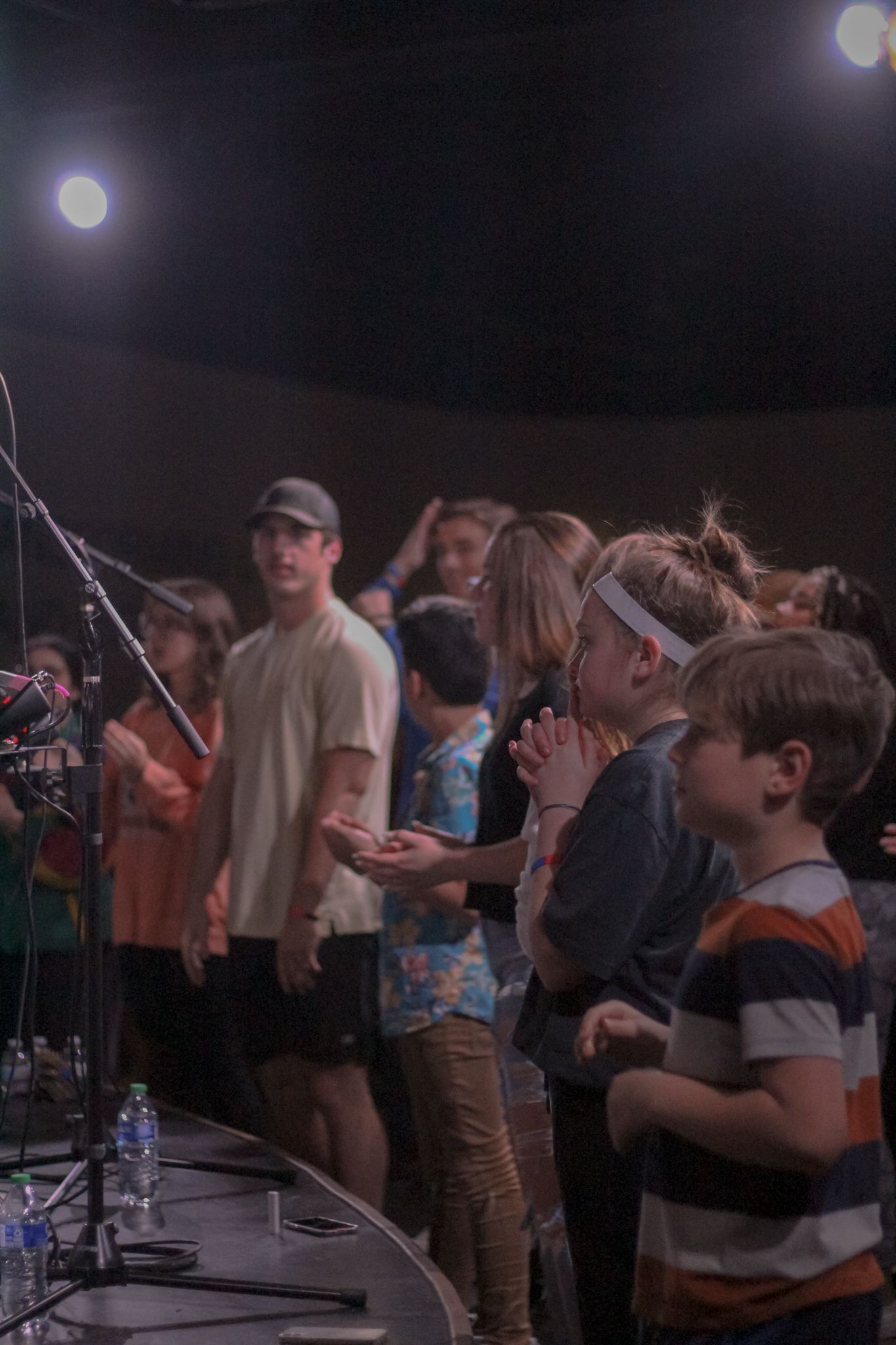 Children and teens gather at the front of a stage during a worship service under stage lights.