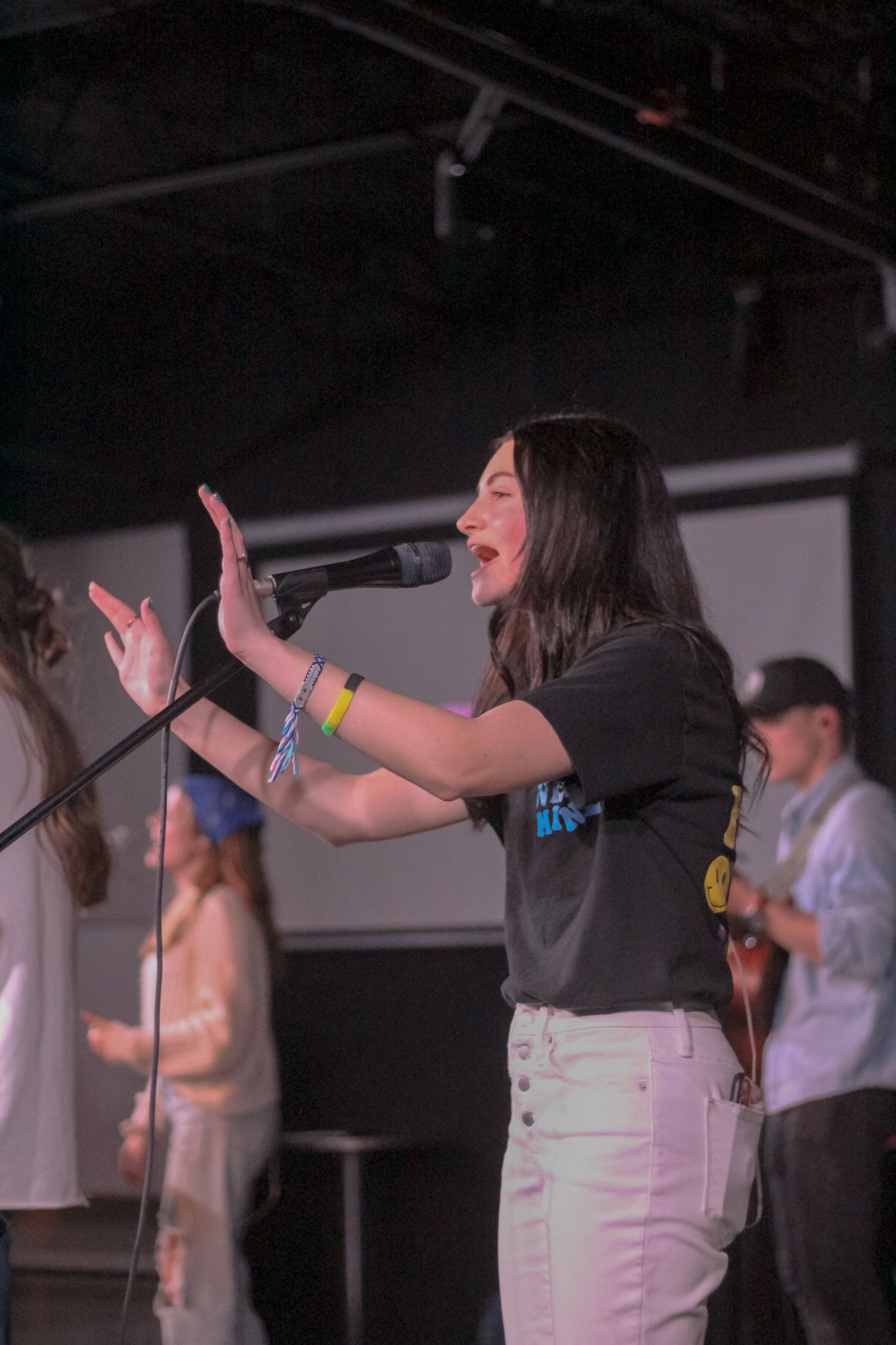 Young woman singing passionately into a microphone on stage wearing a black t-shirt and white jeans.
