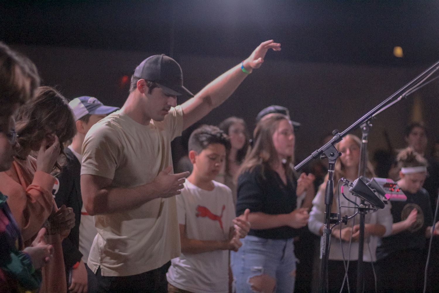 A young man raises his hand in worship while holding a phone during a crowded church service.