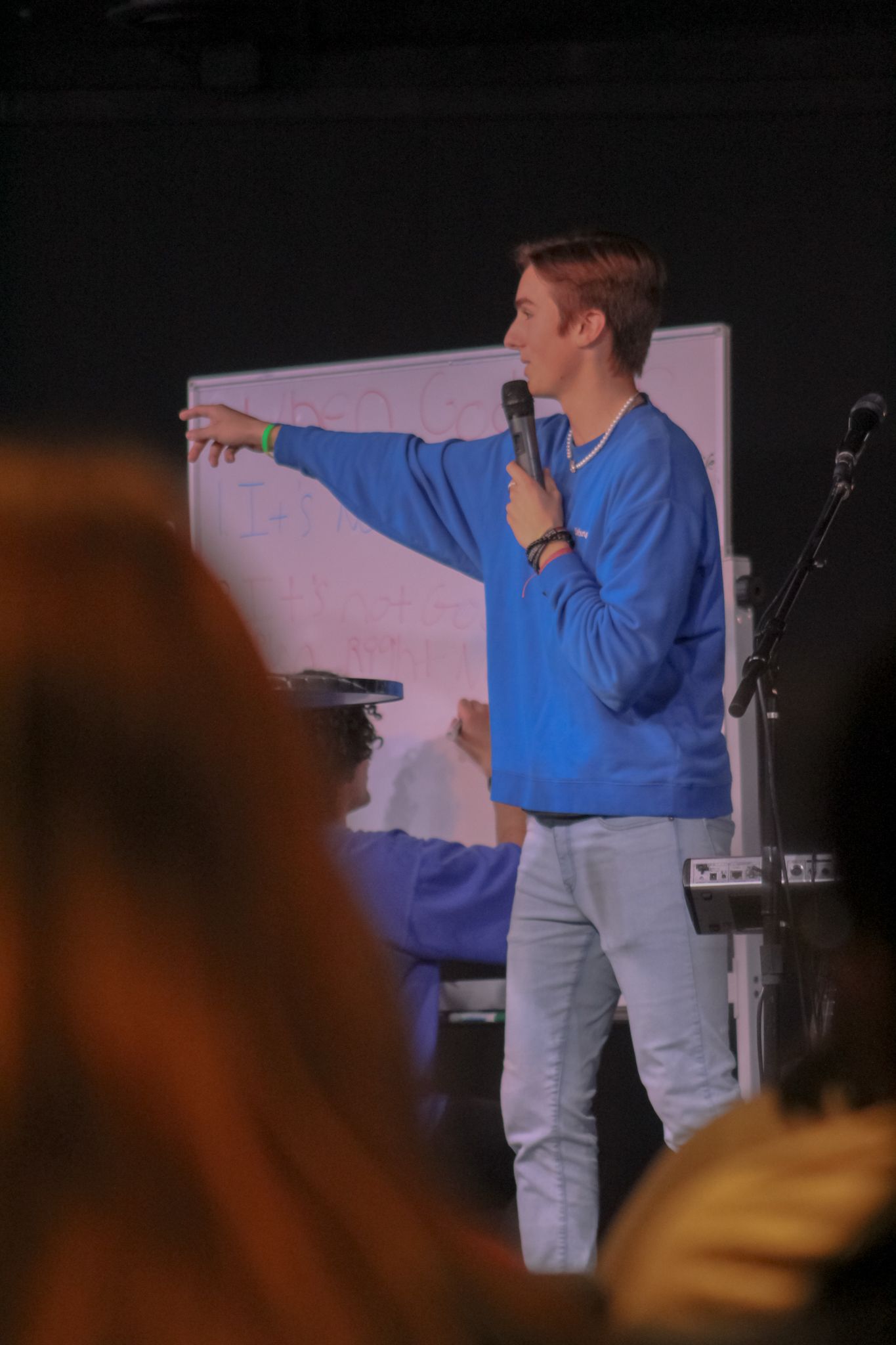 A young man in a blue sweatshirt points to a whiteboard while speaking into a microphone.
