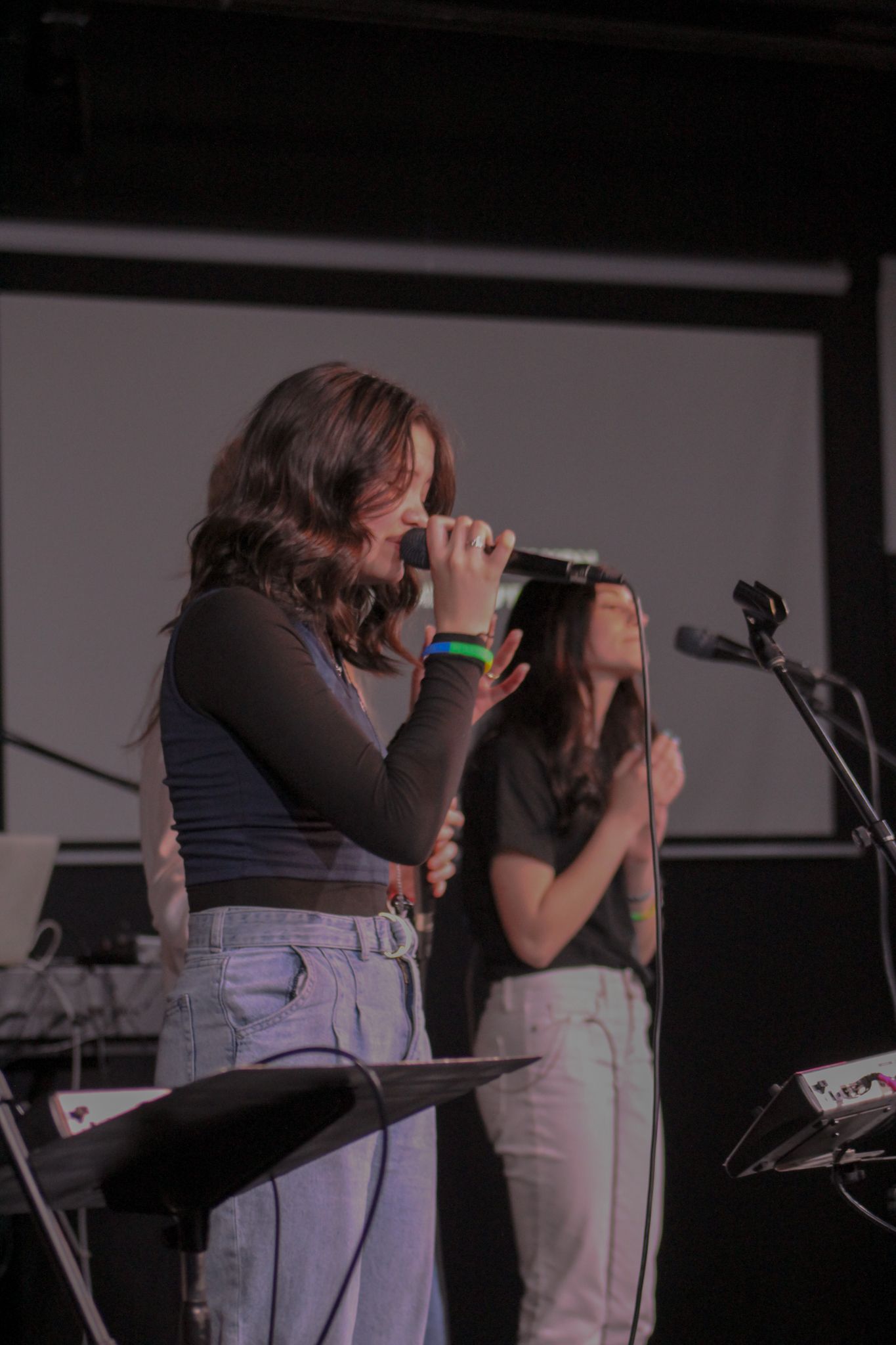 Two young women perform on stage, one singing into a handheld microphone under bright lights.