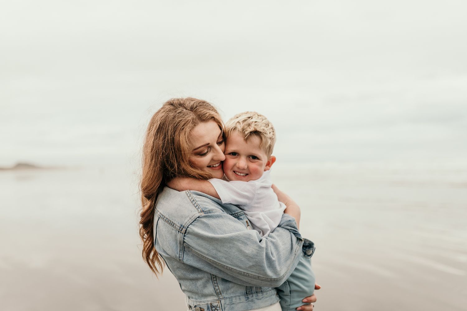 Tender embrace between parent and child on a misty beach in soft, muted tones.