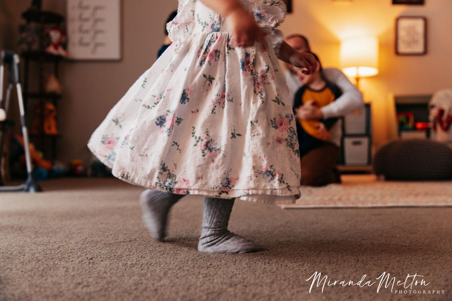 A father wrestles with his kids in the background as a young girl's legs are seen running through the foreground