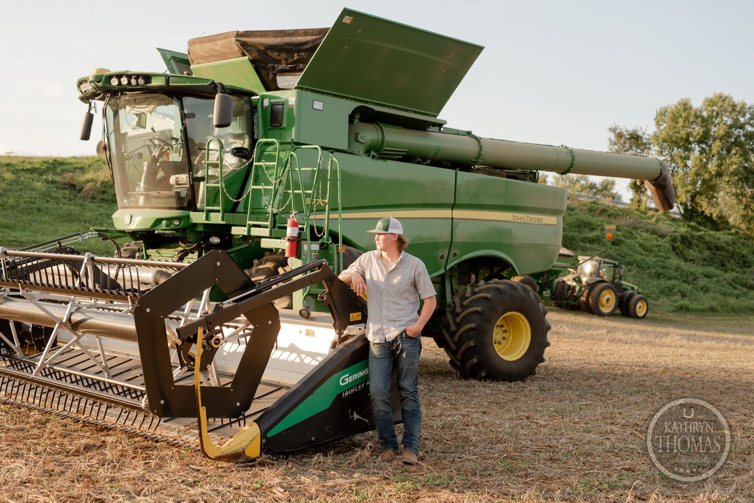 Modern green combine harvester with grain header attachment positioned near storage silos at sunset.