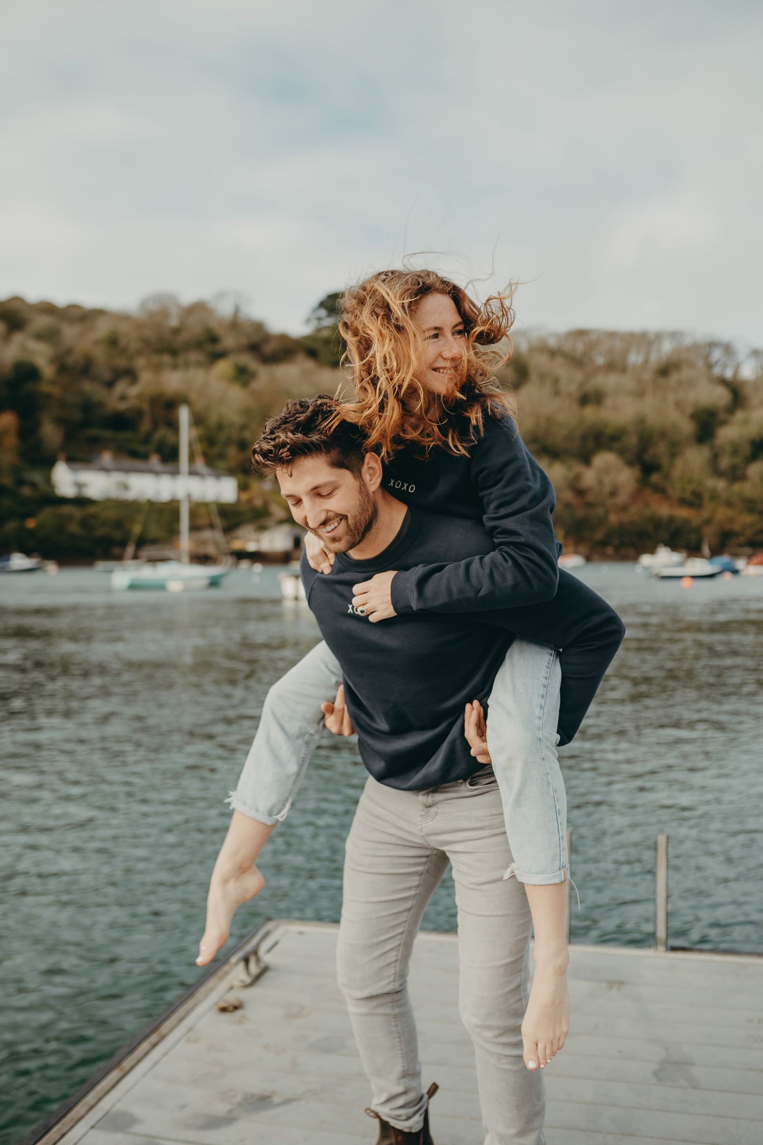 Playful couple enjoying a piggyback ride on wooden dock with boats in harbor.