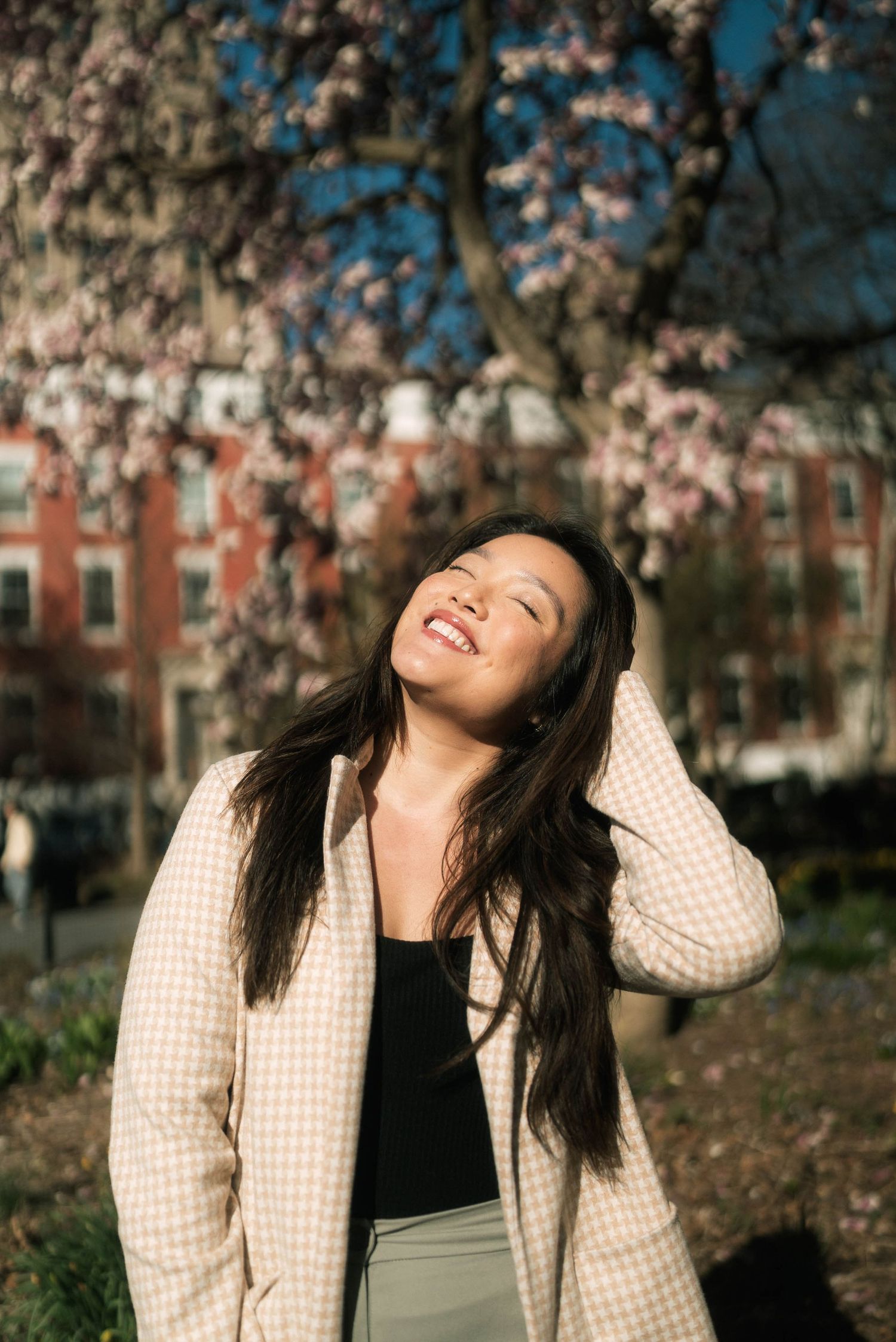 Person wearing a checkered jacket laughing joyfully under cherry blossom trees.
