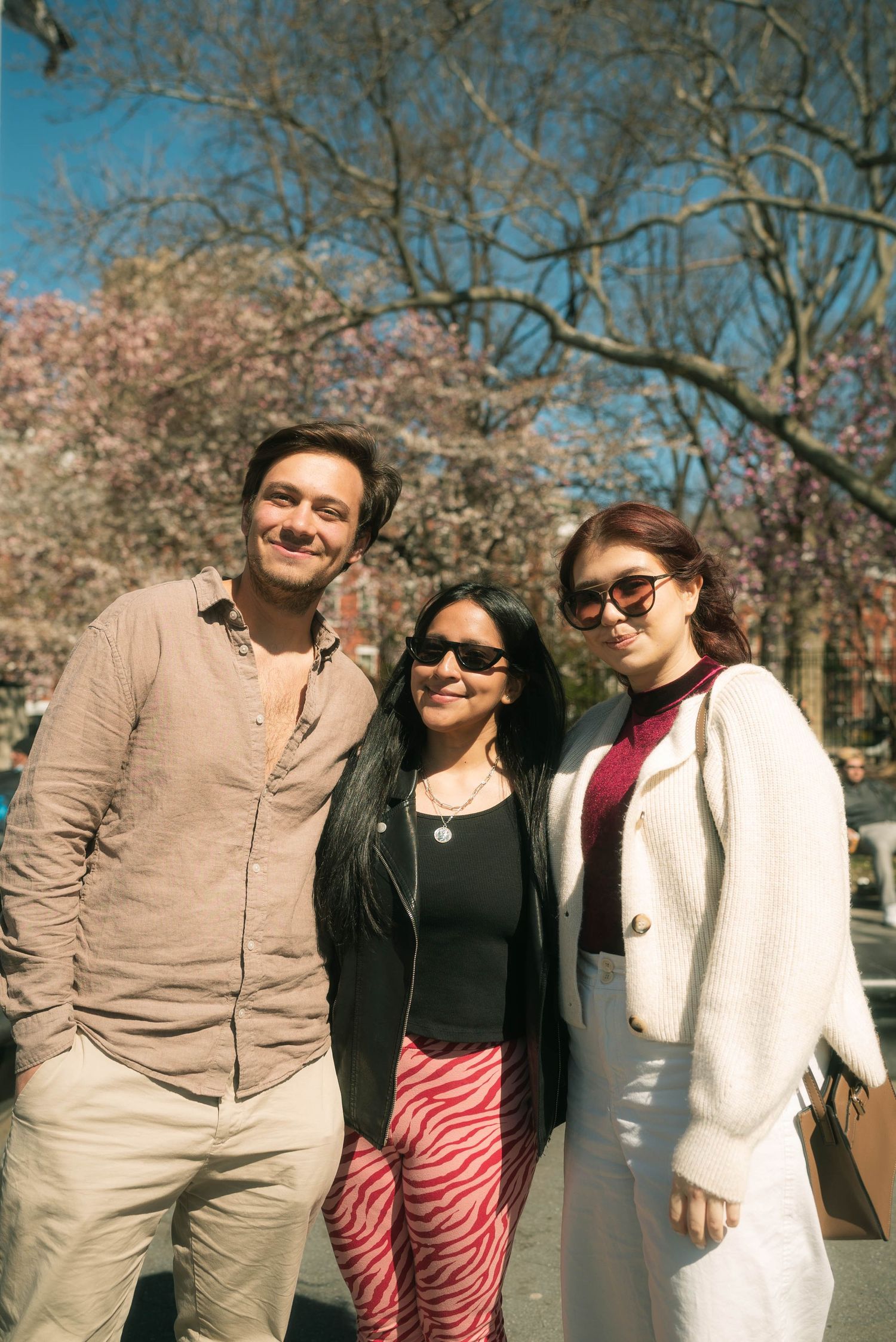 Three friends pose together on a spring day with cherry blossoms blooming behind them.
