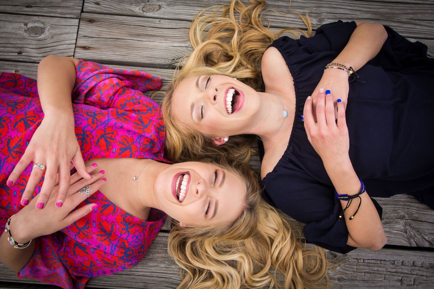 Two people with blonde hair laying down on wooden deck while laughing and wearing pink and black outfits.