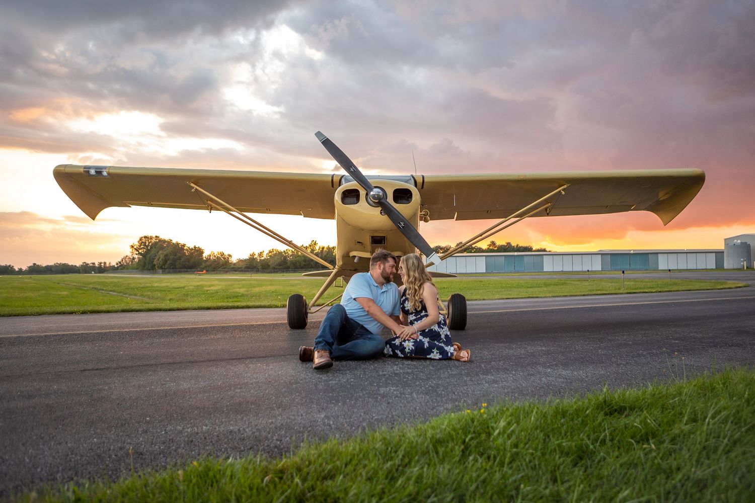 Small yellow airplane parked on runway at sunset with people sitting near its wheels.