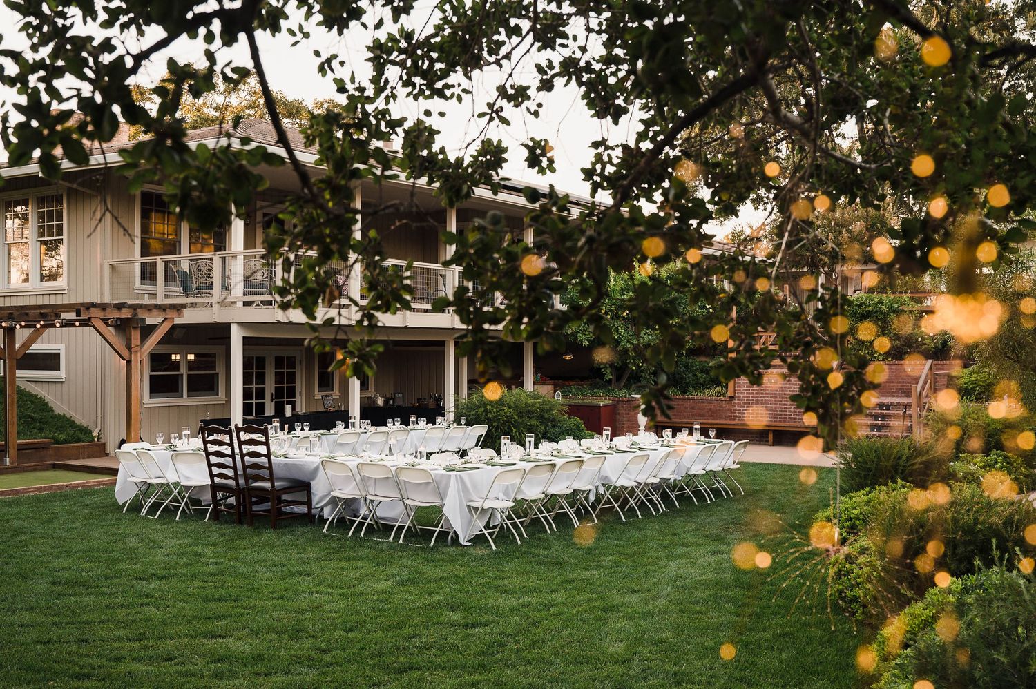 Outdoor wedding reception setup with white chairs and tables on a lawn beside a rustic venue.