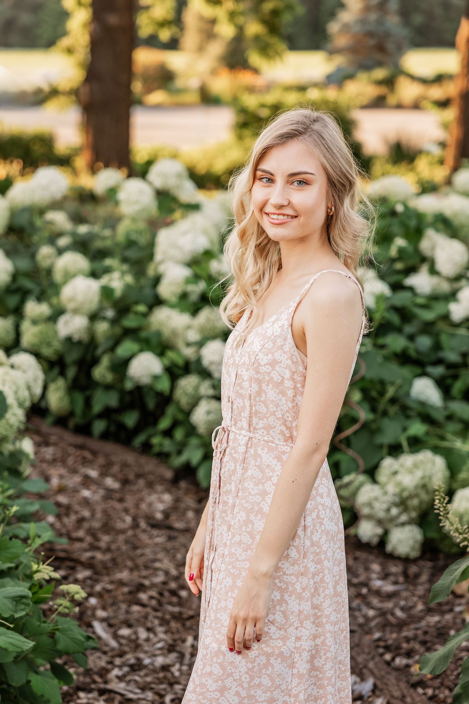 A young woman in a blush pink dress poses in a garden with white hydrangea bushes in the background.