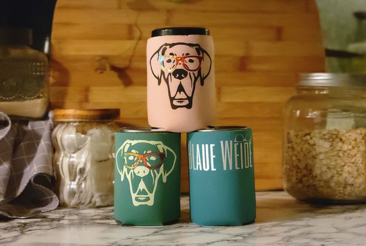 Three dog-themed drink koozies on kitchen counter, featuring Great Dane artwork in pink, green colors.