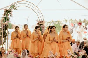 Bridesmaids in matching peach-colored dresses stand under a floral arch during a wedding ceremony.