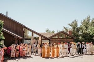 Large wedding party gathered outside rustic wooden venue with colorful traditional attire.