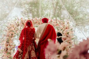Couple in traditional red wedding attire standing beneath floral archway during ceremony.