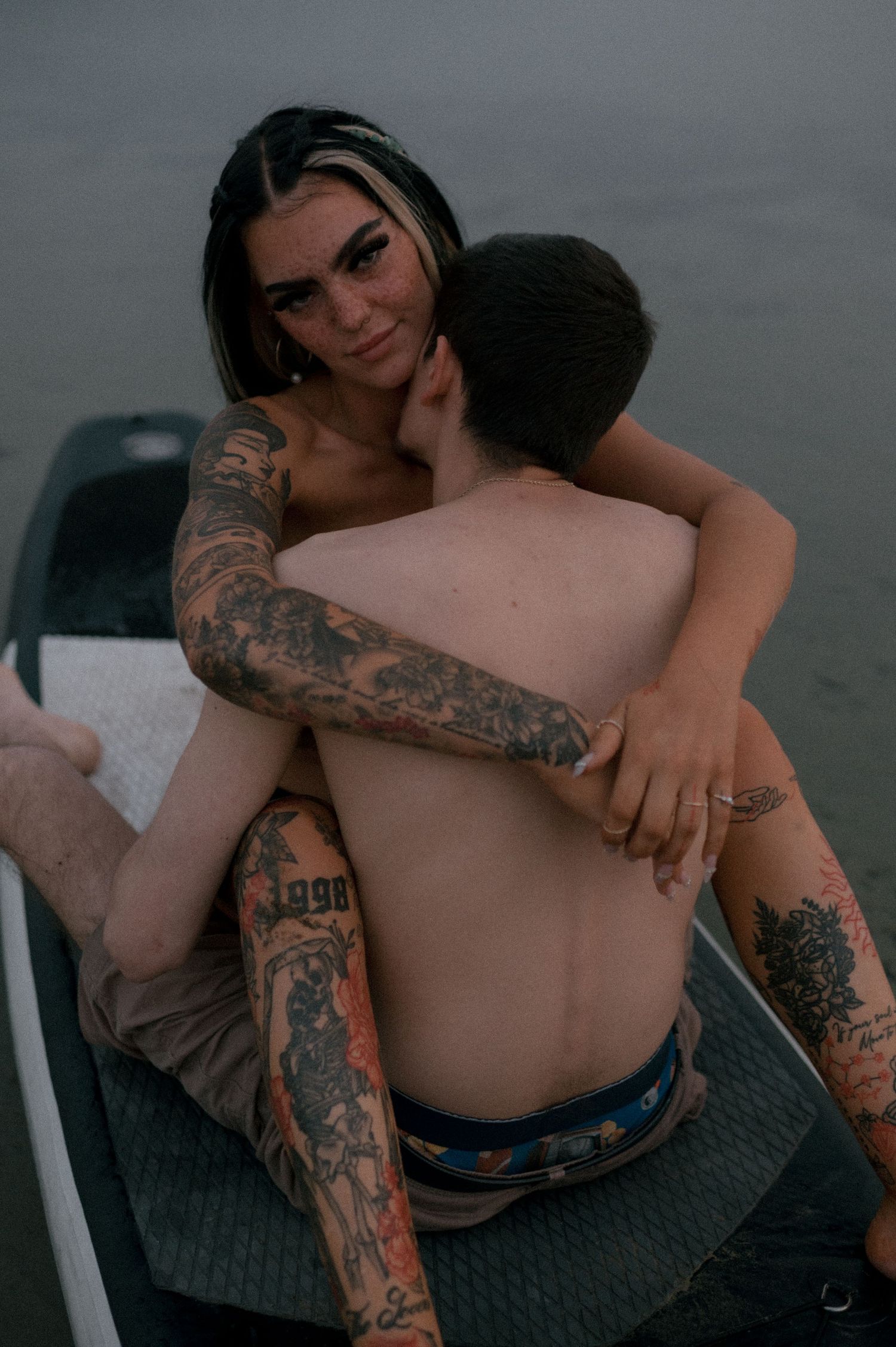 Intimate couple with tattoos embracing on boat deck at dusk.