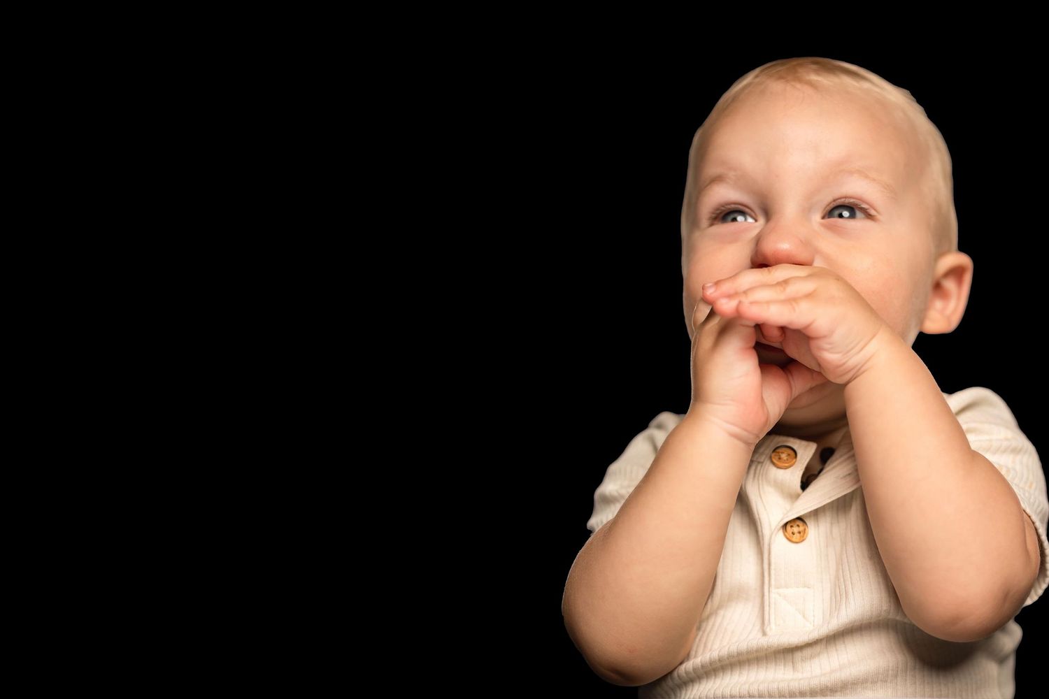 Adorable smiling baby covering mouth with hands against dark black background with copy space.