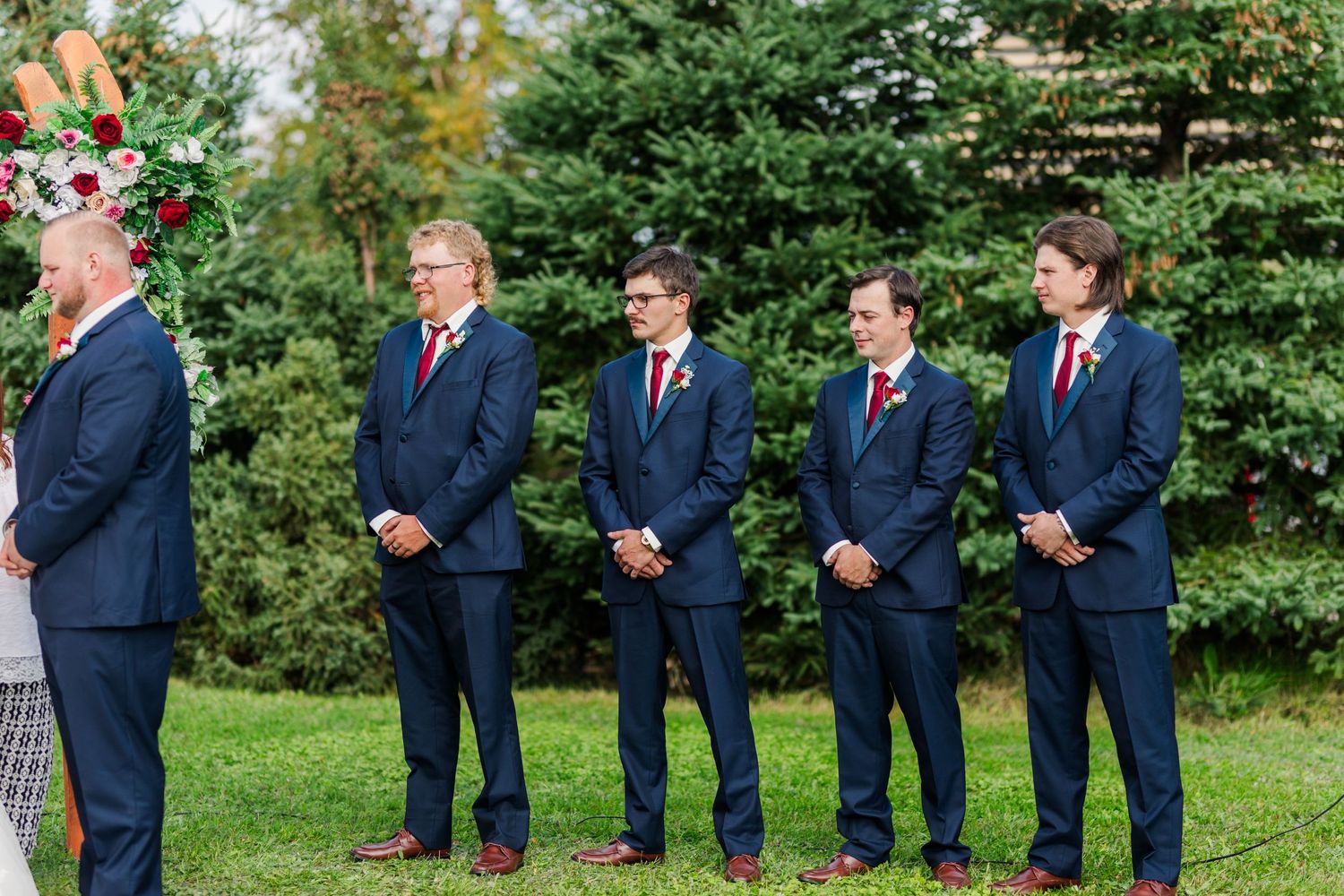Groomsmen in navy suits and red ties stand in line during outdoor wedding ceremony.