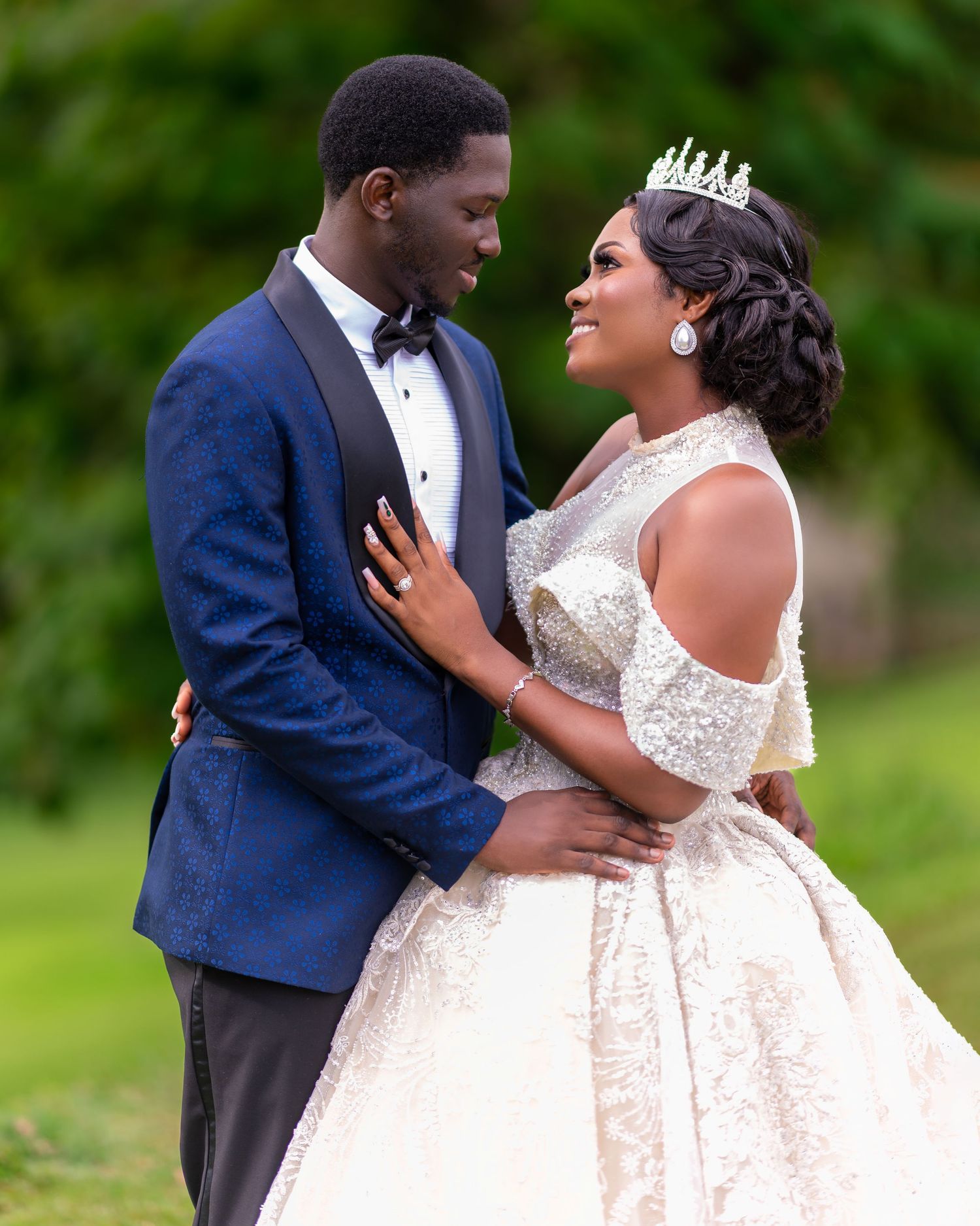 A couple in wedding attire poses outdoors against a lush green garden background.