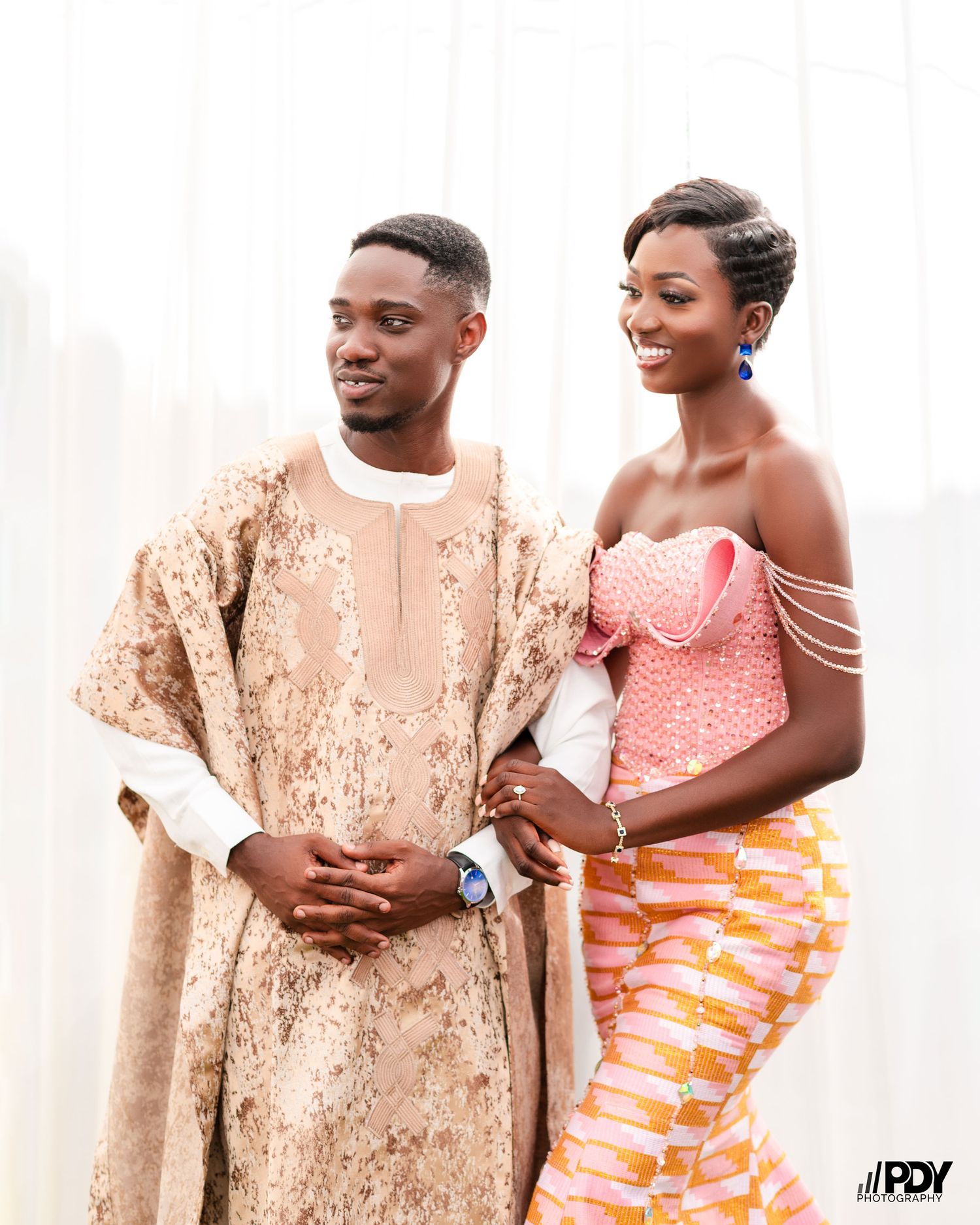 Couple in traditional African attire featuring beige agbada and pink kente-inspired dress.