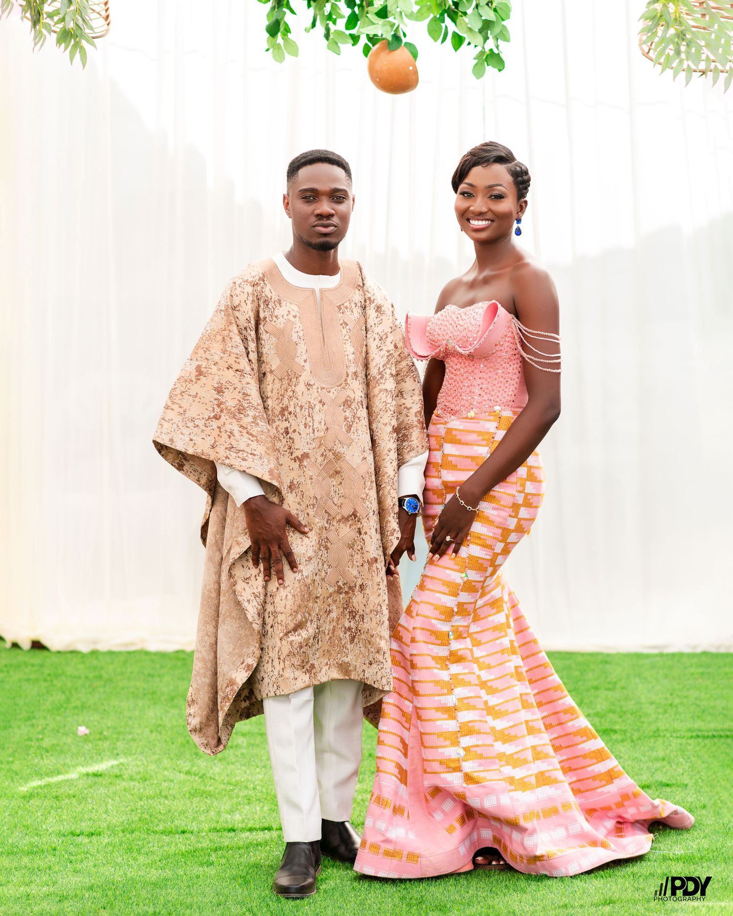 Couple poses outdoors in traditional African wedding attire with decorative orange lanterns.