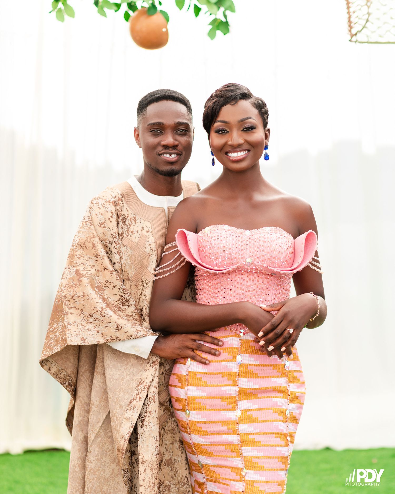 Stylish couple in traditional African formal wear against white backdrop with hanging greenery.