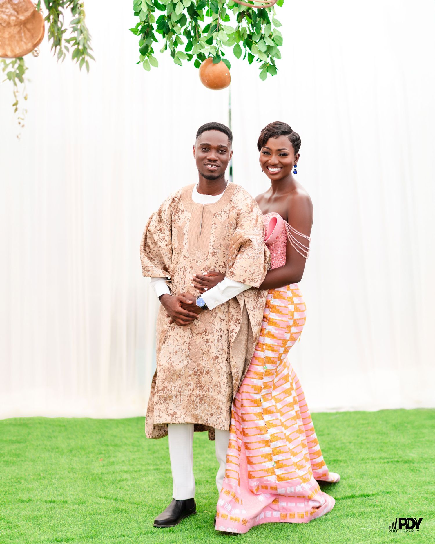 Happy couple in coordinated African formal wear posing on grass with decorative backdrop.