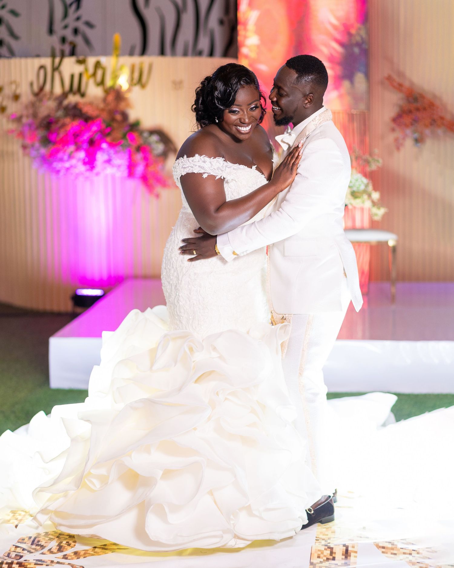 Wedding couple in white attire dancing together under romantic pink and purple lighting.