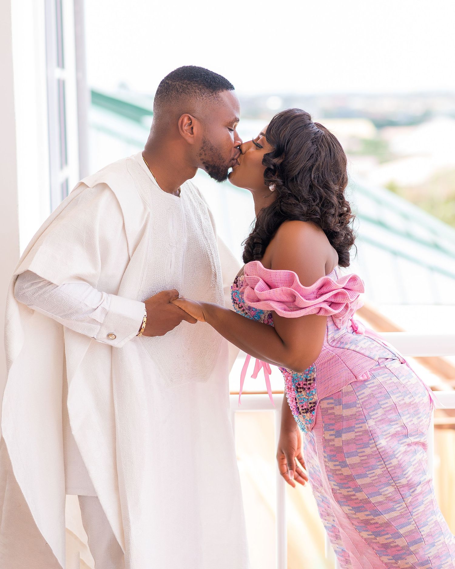 Romantic couple in white and pink outfits sharing playful moments by a window.