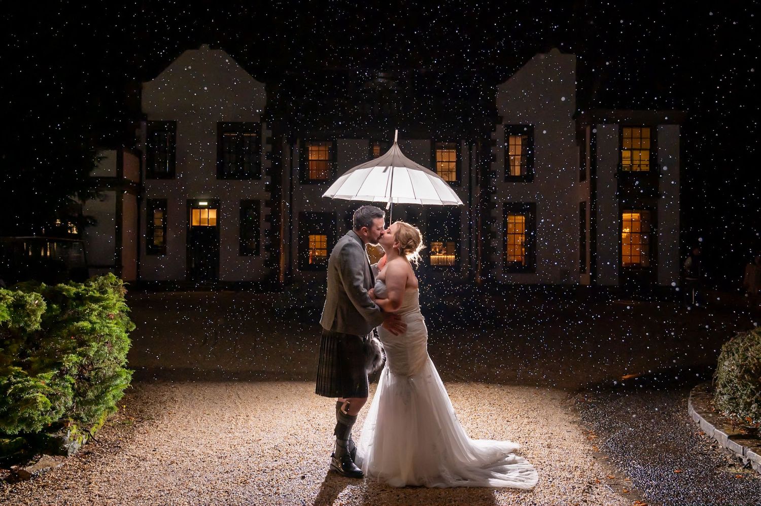 A romantic rainy night wedding portrait under an umbrella with a lit stone building backdrop.
