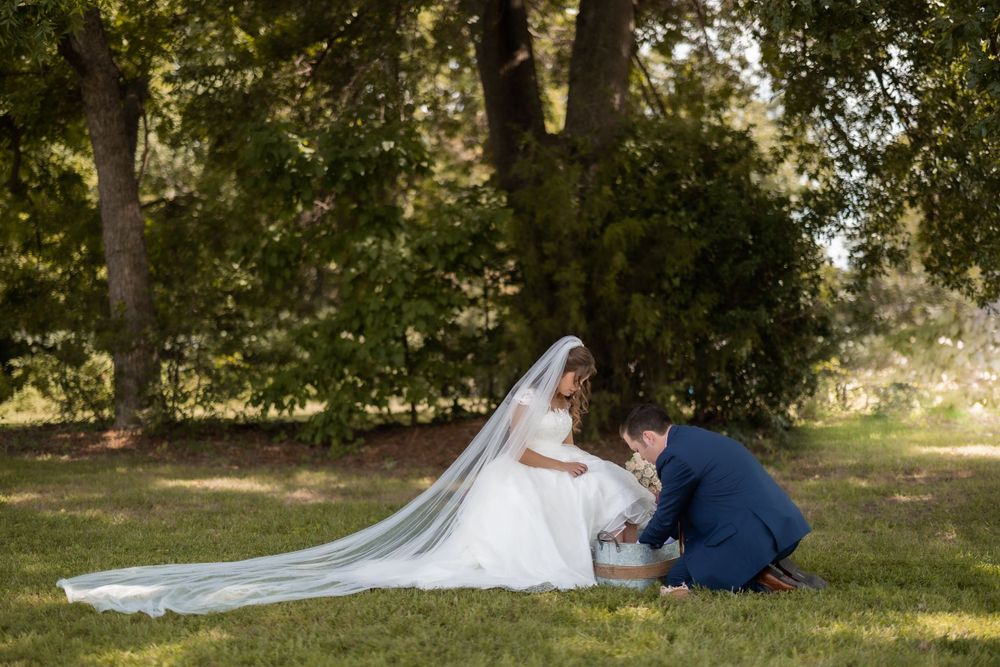 A wedding couple shares an intimate moment on grass under trees with the bride's long veil flowing behind her.
