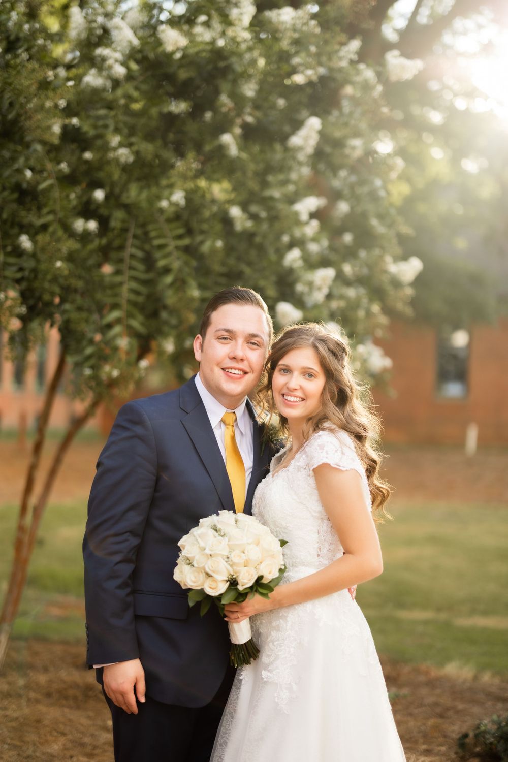 A couple in wedding attire poses outdoors with a bouquet of white roses against a sunlit garden background.