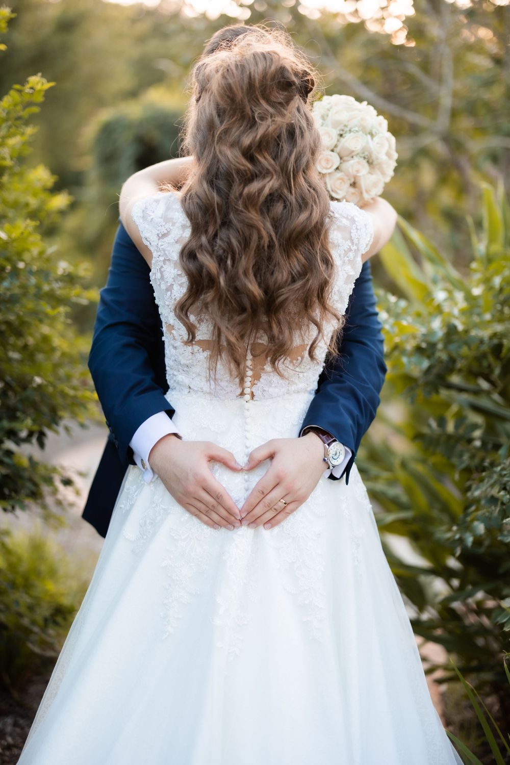 A close-up view of long wavy bridal hair and white wedding dress with hands making heart shape.