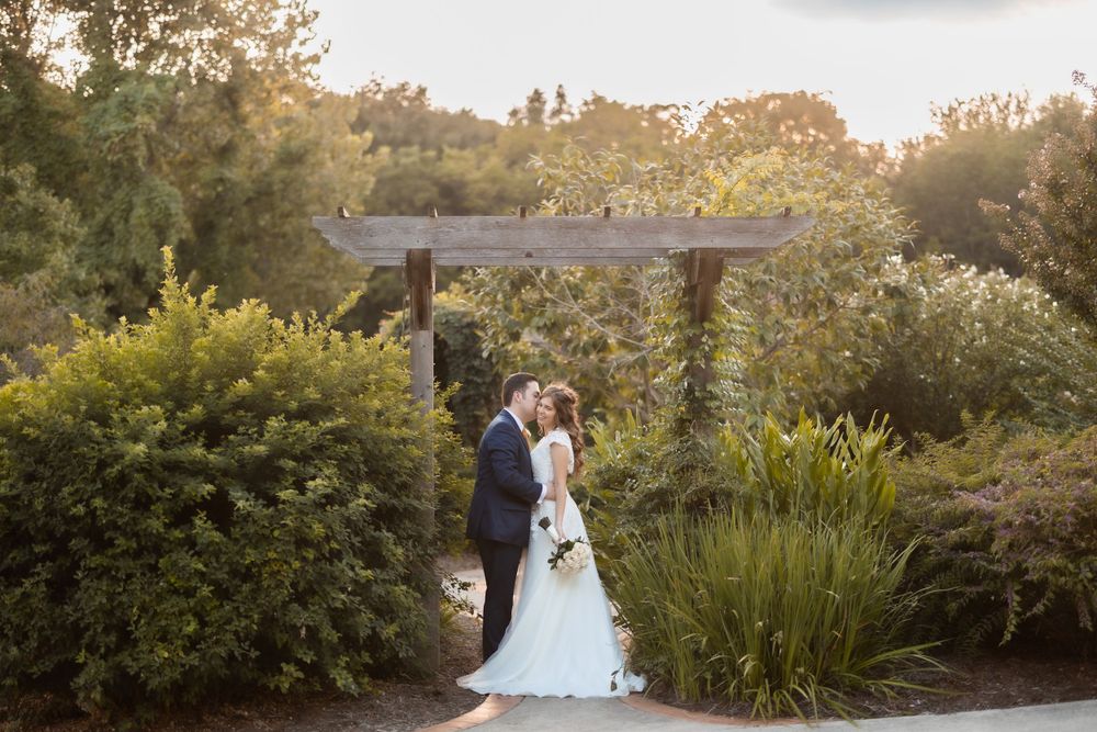 A wedding couple poses under a wooden pergola in a garden setting during sunset.