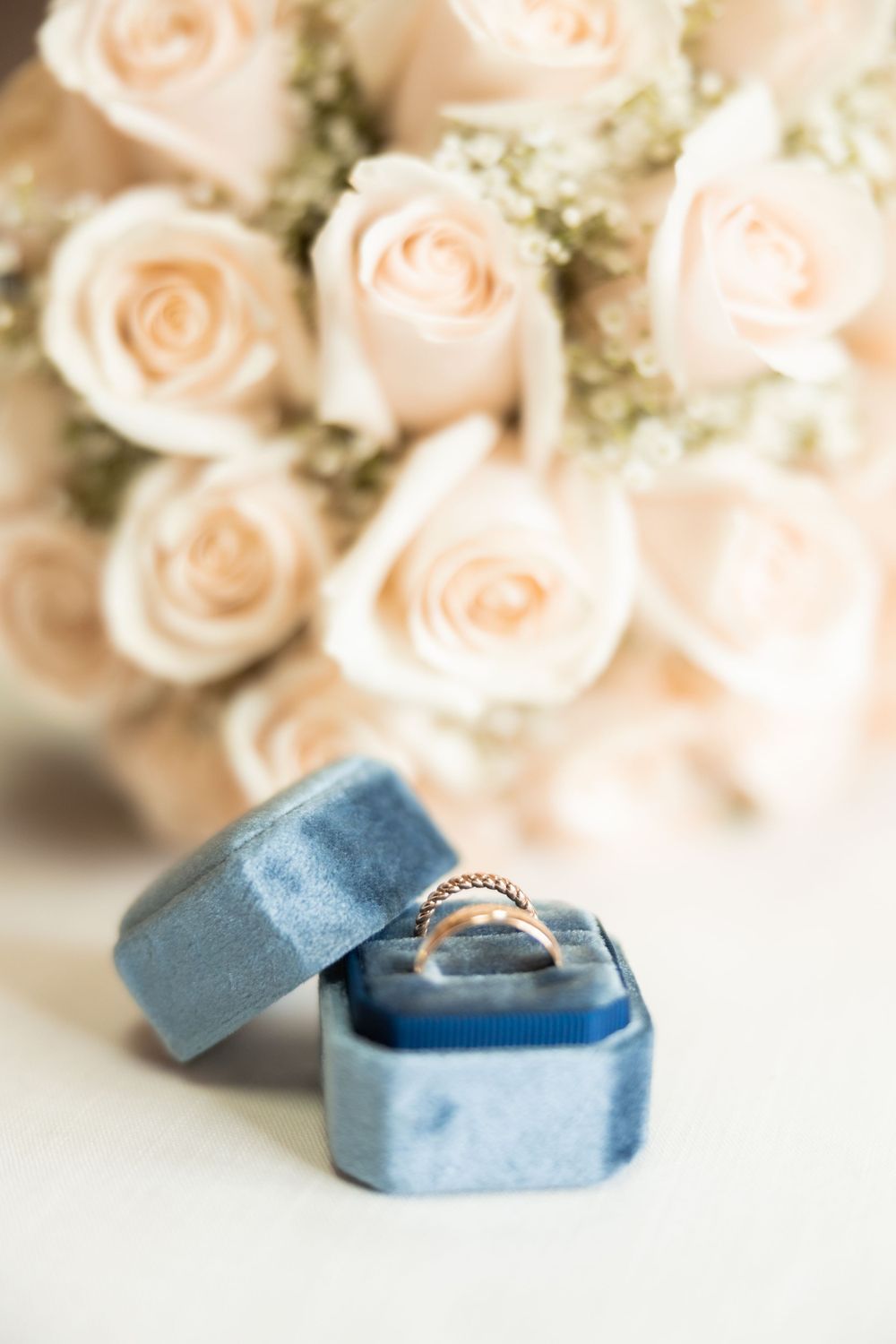 Wedding rings displayed in blue velvet box with soft peach roses in background.