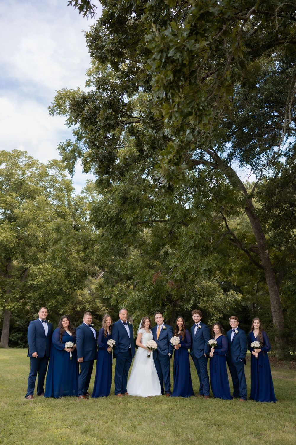 Large wedding party in navy blue attire poses under trees for formal group photo.