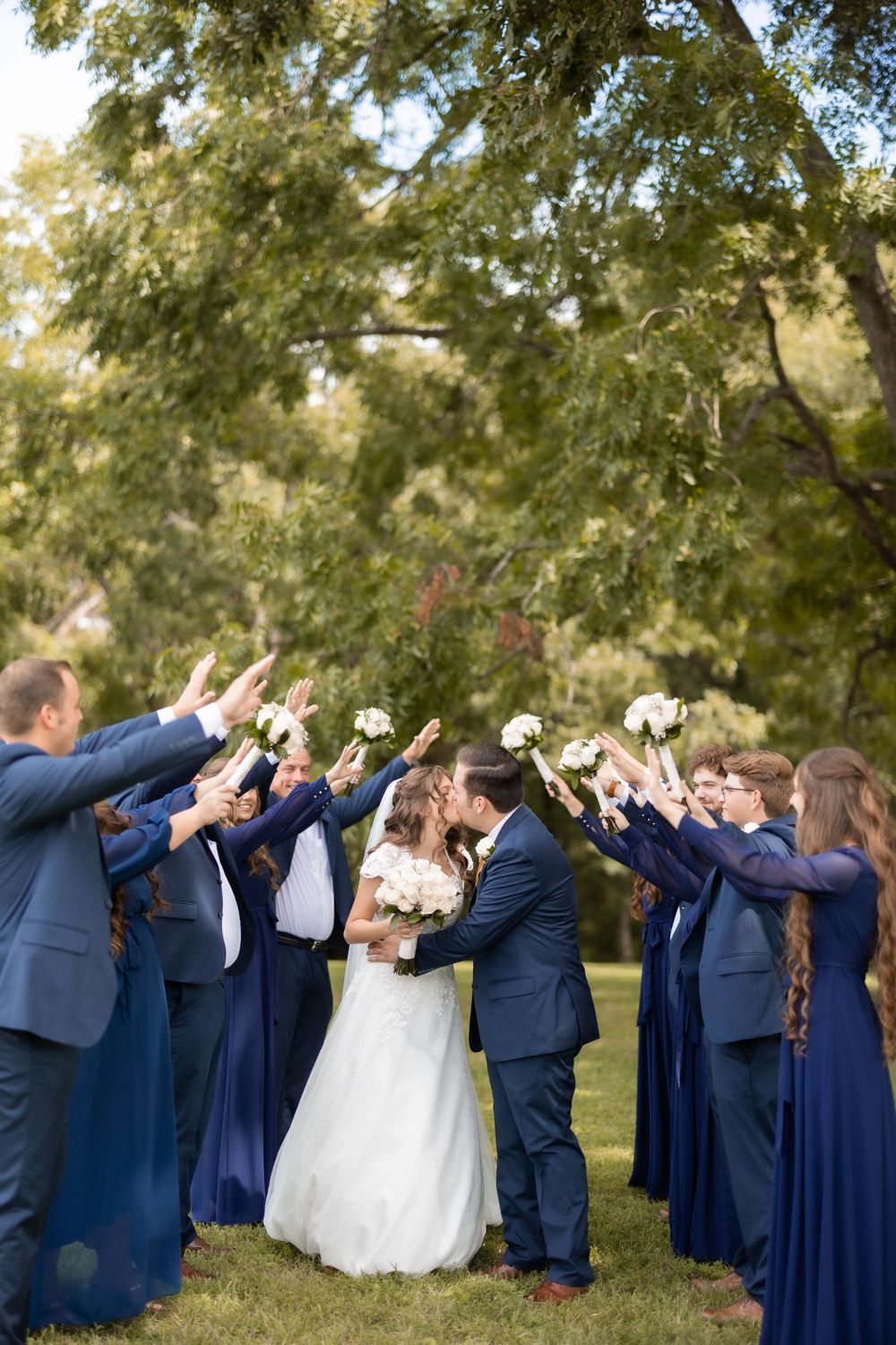 Wedding party in navy blue forms tunnel with raised arms over newlyweds.