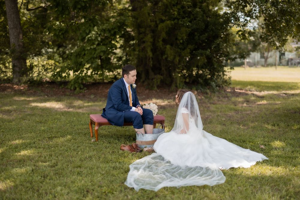 Newlyweds participate in traditional foot washing ceremony on rustic wooden stool outdoors.